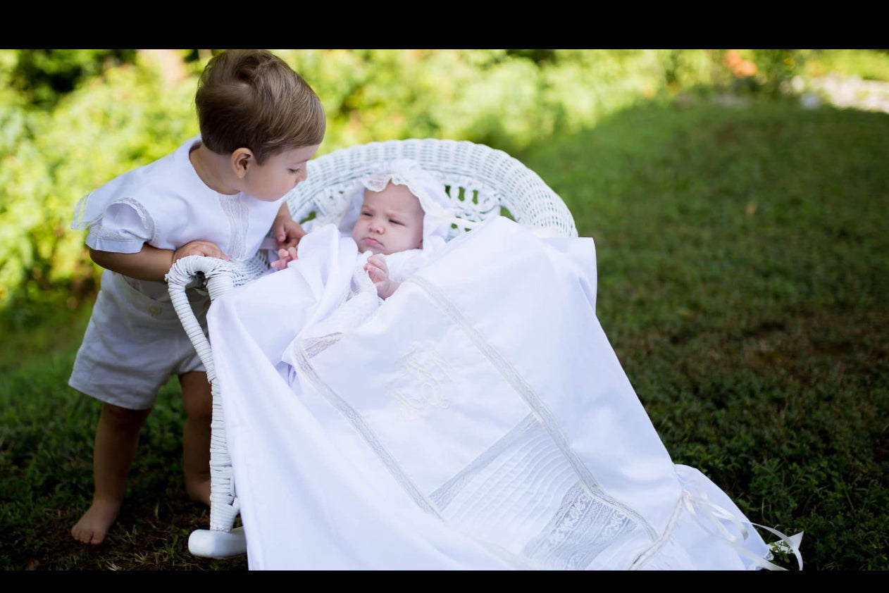 Two children in white heirloom clothing, cotton batiste, French lace outdoors, one holding a baby wrapped in a white blanket.