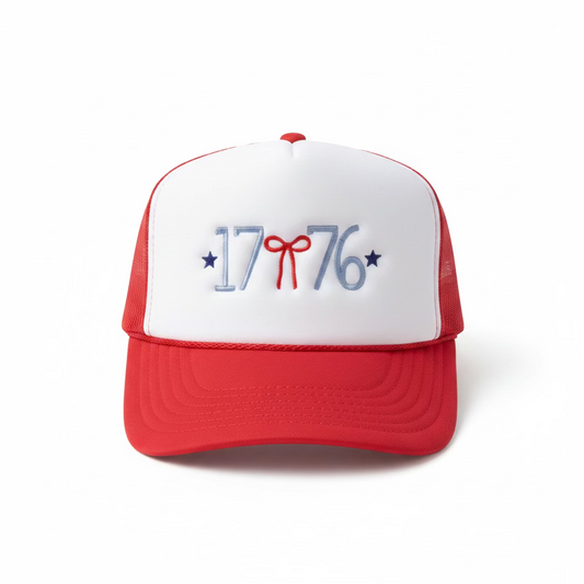 Woman wearing a red and white 1776 embroidered trucker hat outdoors with an American flag in the background — patriotic Fourth of July hat with red bow and blue stars