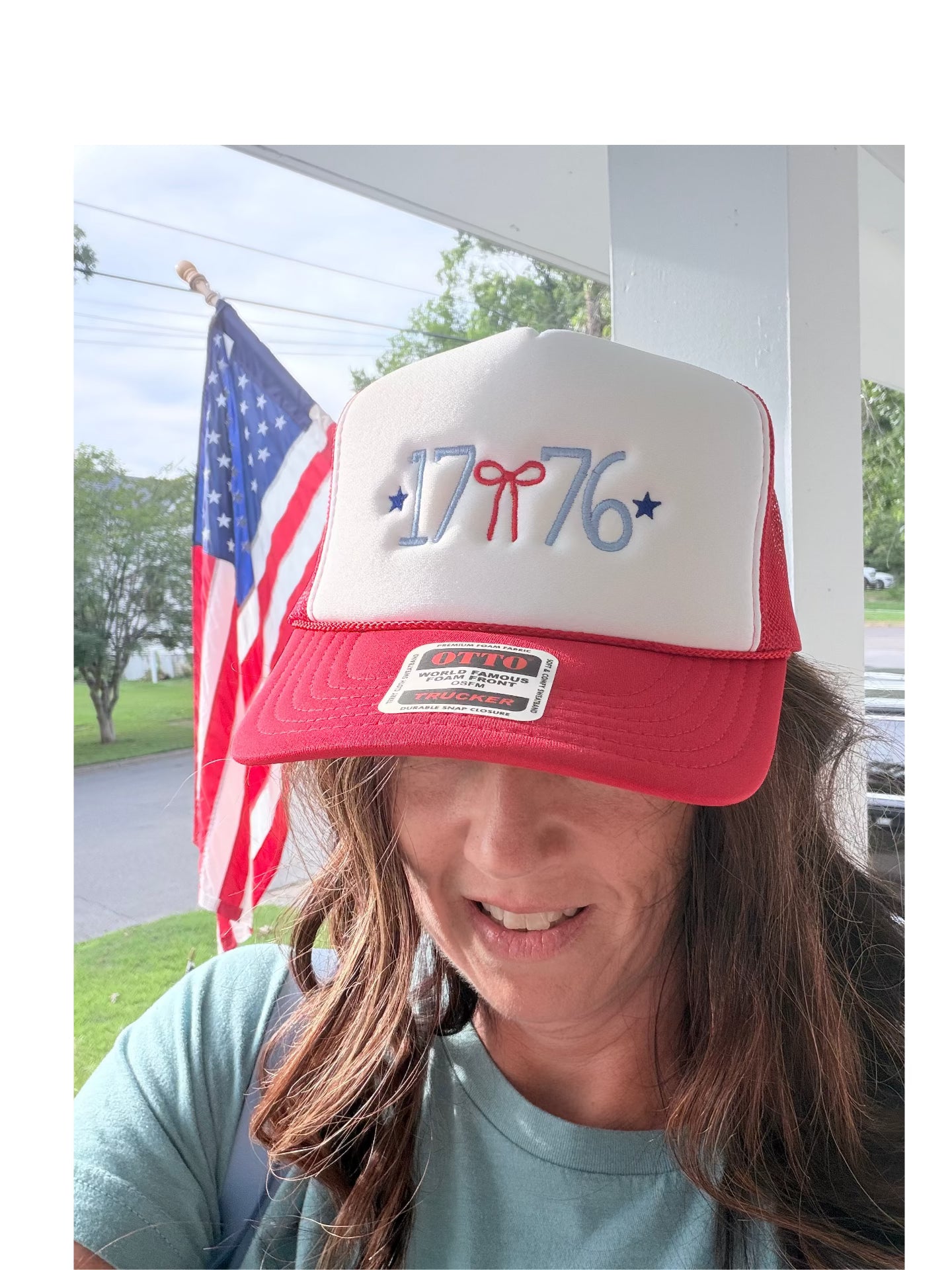 Woman wearing a red and white 1776 embroidered trucker hat outdoors with an American flag in the background — patriotic Fourth of July hat with red bow and blue stars