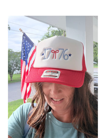 Woman wearing a red and white 1776 embroidered trucker hat outdoors with an American flag in the background — patriotic Fourth of July hat with red bow and blue stars