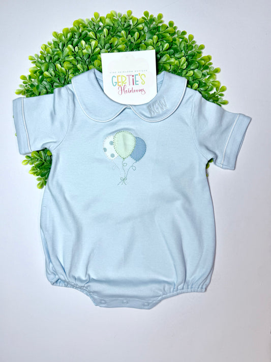 A light blue baby short-sleeve romper with a Peter Pan collar, featuring an embroidered balloon design in mint green and light blue. The romper is displayed flat on a white background, styled with a green artificial hedge and a paper brand tag that reads 'Gertie's Gardens'.
